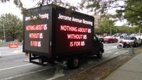 A black box truck is parked along the curb under a tree. The truck's large LED screens read, "Jerome Avenue Rezoning. Nothing about us without us is for us"