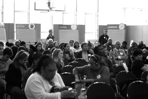 Attendees fill seats on the floor of a gymnasium, with multiple poster displays in the back of the room that read "Eating," "Living", and "Learning"