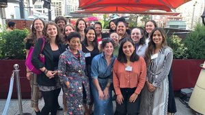 A group of female presenting folks gathered on a rooftop patio smiles for the camera.