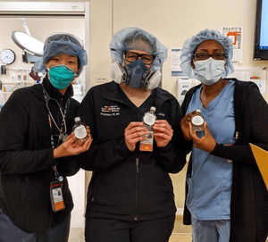 Three healthcare workers wearing masks and protective equipment pose with miniature bottles of hand sanitizer