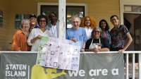 A diverse group of people stands smiling on a porch holding a large plan