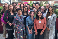 A group photo of smiling female-presenting people standing together on a rooftop with lots of greenery.