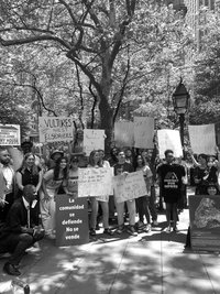 A black and white photo of a group of activists in a park, holding up signs that say, 'Vultures Nest Elsewhere,' 'La comunidad se defiende, no se vende,' 'East New York is Under Attack - What do we do? Stand Up and Fight Back!', and 'Housing Justice Now', among others. A woman holds a shirt that reads 'Protect Our Homes,' while another sign shows a map labeled 'Homes Flipped in NYC'.