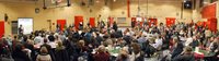 A school gymnasium filled with people sitting around tables while a speaker holding a microphone addresses the room.